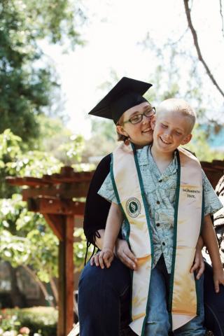 A young woman wearing a black cap and gown with a gold honor stole with green trim partially hugs a smiling young boy standing in front of her.