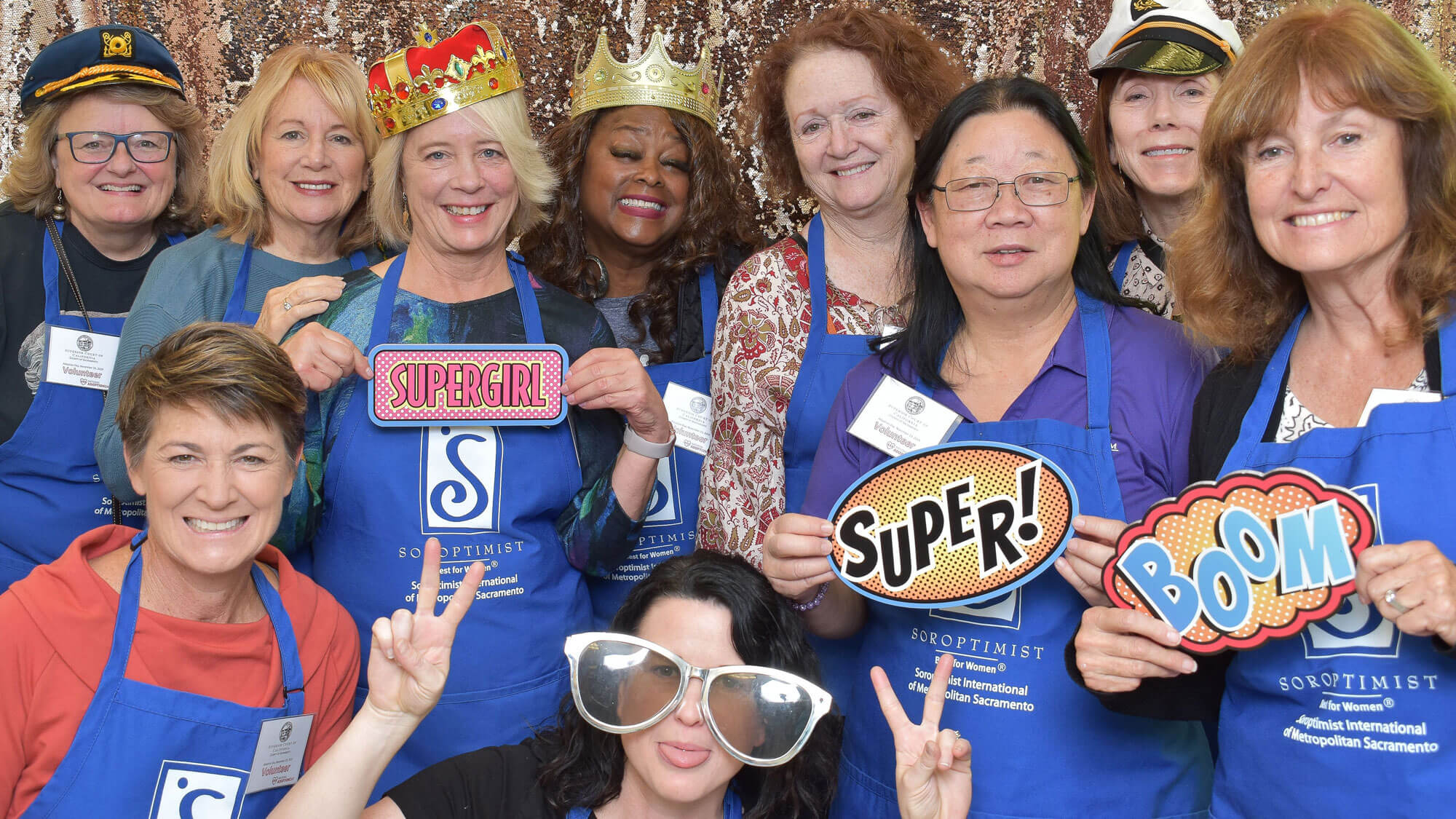 Close-up of a group of middle-aged women wearing blue Soroptimist aprons, all smiling at the camera while some hold up comic-style signs that say "Supergirl", "Super" and "Boom".
