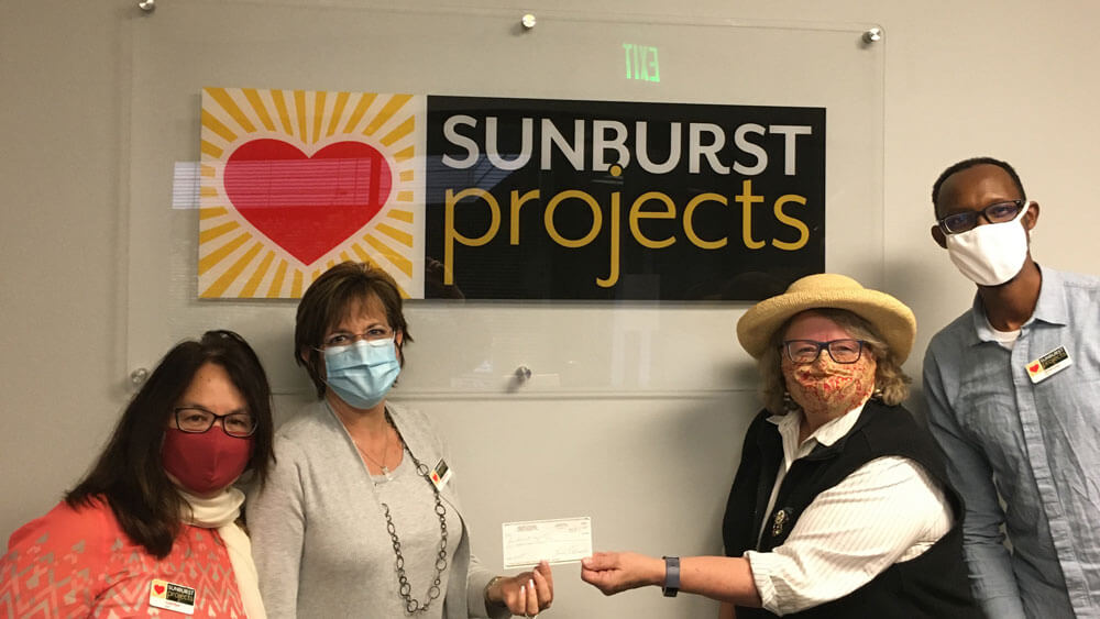 Three women and one man, wearing cloth and sterile face masks, looking at the camera while standing in front of large Sunburst Projects logo encased in glass.
