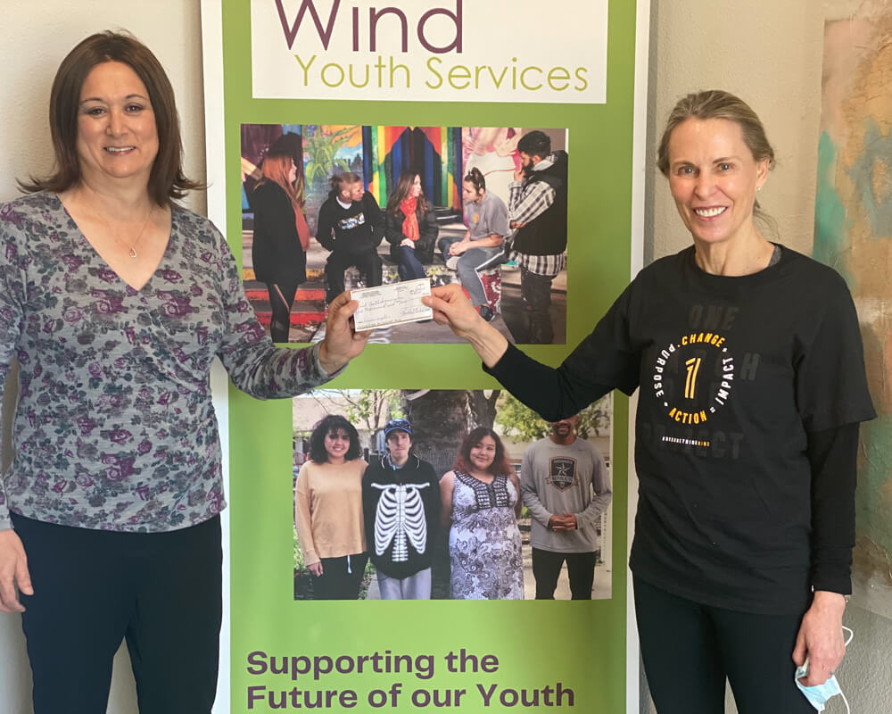 Two women standing on either side of the front of a light green vertical banner that says, "Wind Youth Services" while each holding one end of a check.