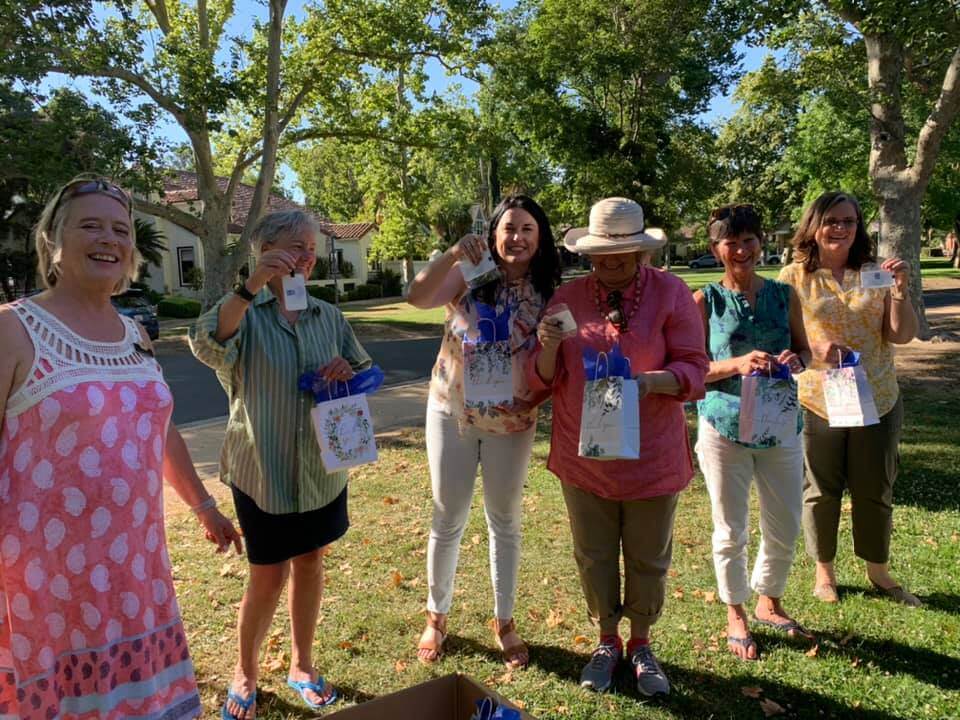 Group shot of six Soroptimist members standing on a front lawn, some holding gift bags and holding up items from the bags.