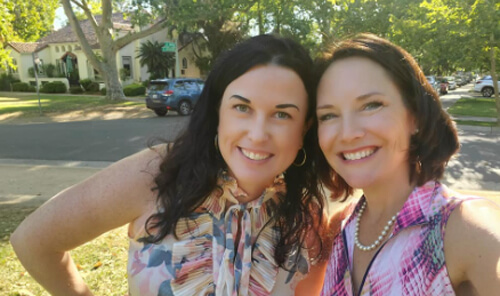 Close-up of two brunette females standing closely together and smiling at the camera, with a view of a suburban neighborhood in the background.