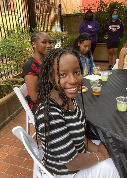 Close-up of young black women sitting at a table outdoors. The young woman in the foreground is wearing a black, gray, and white striped top with, has long braids and is smiling at the camera.