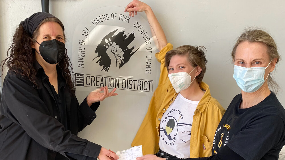 Close-up of three women standing facing the camera while all holding a clear sign that says, "The Creation District" and holding a check.