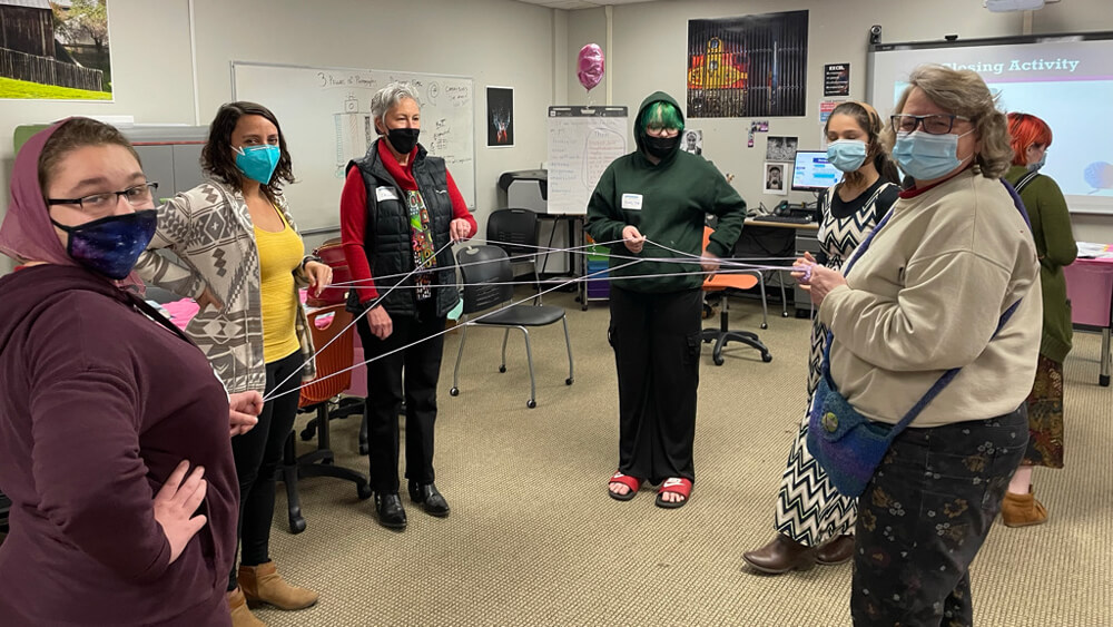 Group of women and teenage girls standing in a classroom in a circle, each holding part of a long string that forms a web.