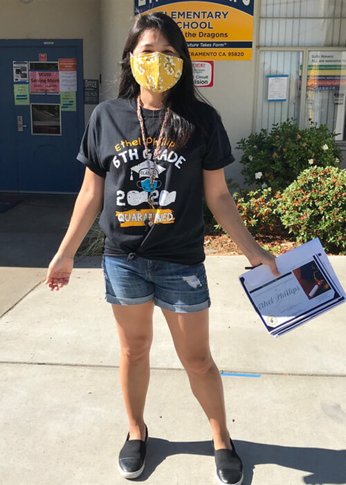 A young girl standing in front of an elementary school, wearing a black t-shirt, denim jean shorts, and sneakers, and holding a certificate in her hand.