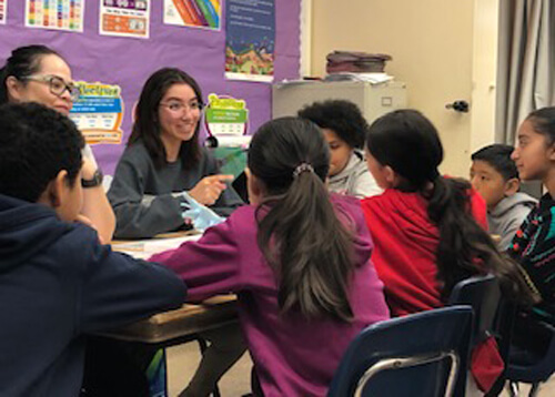 Group of students and teachers gathered around a classroom table, smiling and in the middle of a discussion.