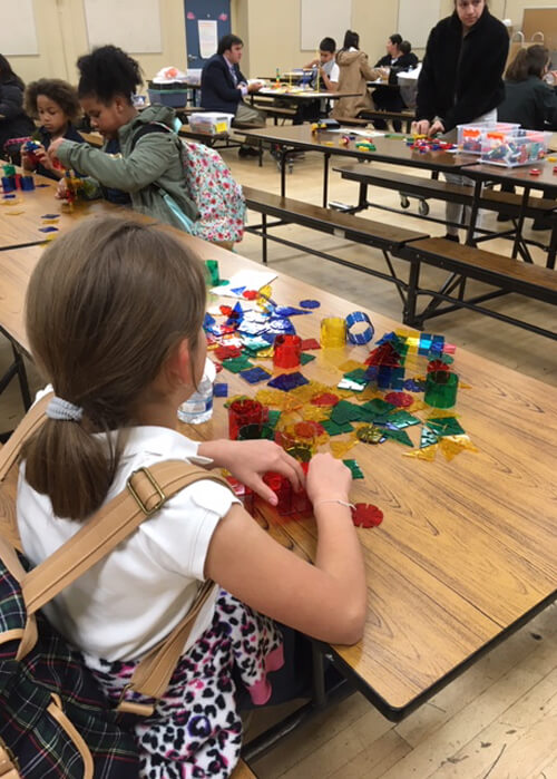 Various young students and adults sitting a different tables in a cafeteria together, working on different creative projects. The girl in the foreground has her back turned to the camera, is wearing a plaid backpack, and is sitting by herself, playing with various plastic shapes.