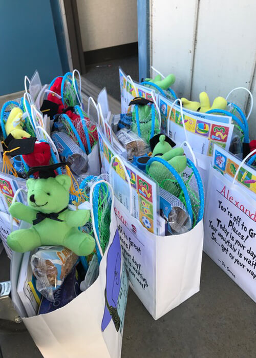 A group of white goodie bags on the floor filled with an assortment of items, including colorful plush teddy bears wearing graduation caps and snacks.