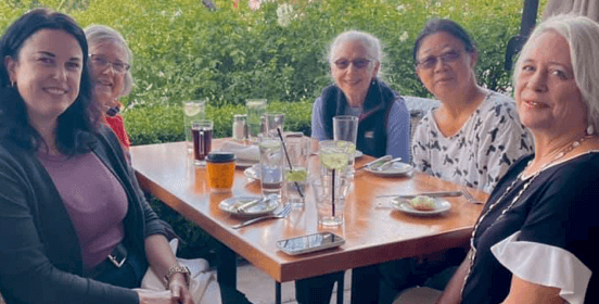 Five women of different ethnicities sitting together at a table during a meal, all dressed in nice but casual attire.