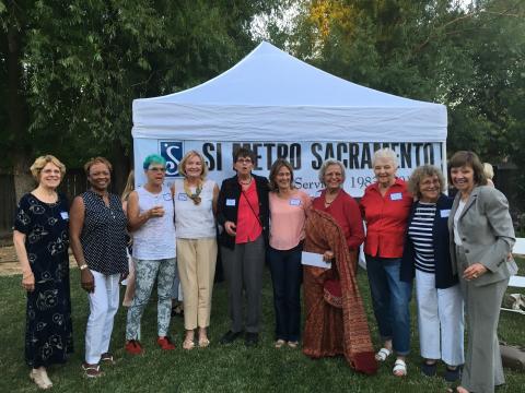 Wide shot of a group of 10 women standing in a row next to each other in front of a white tent with a large banner that says, "SI Metro Sacramento".