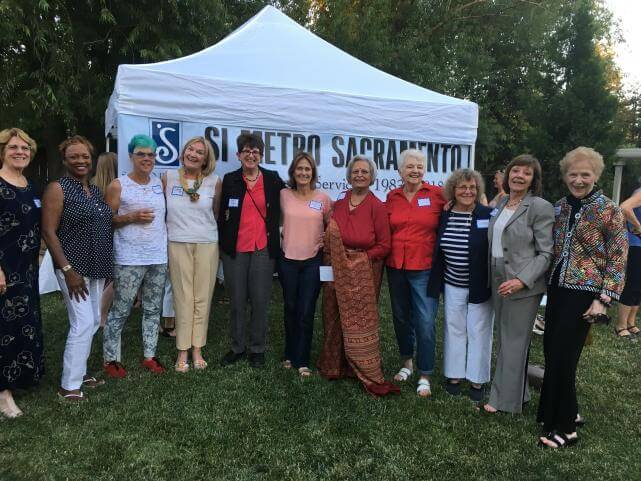 A group of older women of various races stand next to each other outdoors in front of a white tent and a banner that says, "SI Metro Sacramento".