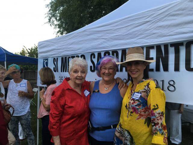 Three older women stand together, the one in the center has her arms wrapped around the others' shoulders. A banner hangs on the white tent behind them, which says, "SI Metro Sacramento".