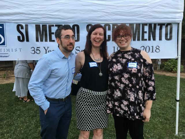 A young man with a mustache, beard, and glasses, stands close to two young women, both dressed in dark summery outfits, in front of a white outdoor tent and a banner that says, "SI Metro Sacramento".