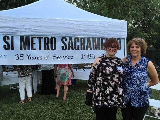 A young woman wearing a floral print top standing next to an older woman wearing a blue leopard-print top, in front of a white outdoor tent and a banner that reads, "SI Metro Sacramento".