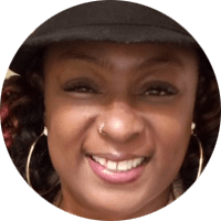 Close-up of a black woman's face. She is smiling, has a nose piercing, wearing a black cap, and large hoop earrings.