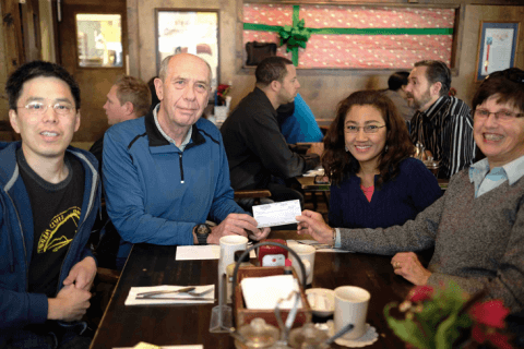 Two men and two women sit at a table in a restaurant together, the older man and older woman are each holding up a corner of a check, and all are smiling at the camera.