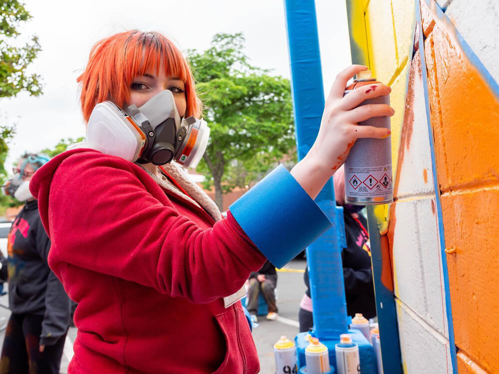 A teenage girl with short orange hair looks wears a protective mask and looks at the camera while she holds a can of spray paint to an outdoor brick wall.