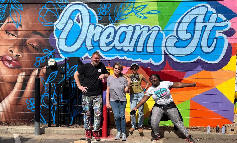 A man and three young women of different races stand in front of a colorfully-painted mural featuring the image of a young black woman and the words, "Dream It".