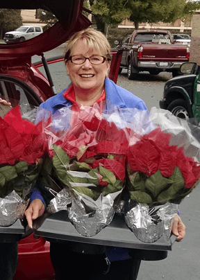 A middle-aged caucasian woman wearing dark-rimmed glasses standing by the open trunk of a red car while holding a tray of poinsettias and smiling.