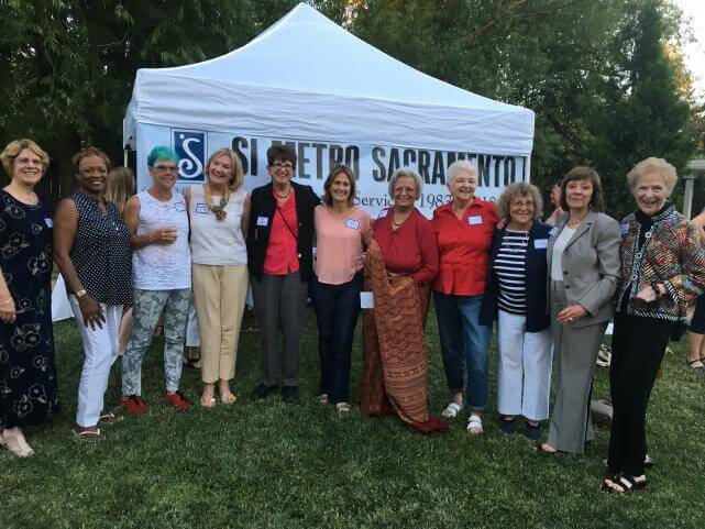 A group of older women of various races stand next to each other outdoors in front of a white tent and a banner that says, "SI Metro Sacramento".