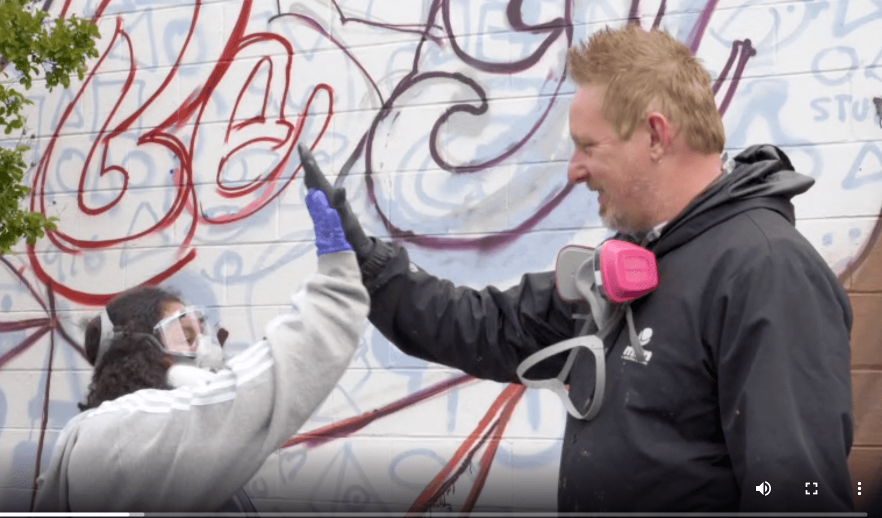 Teenage girl and a man both wearing protective masks for painting high-five each other while standing in front of a brick wall featuring the beginnings of mural art.