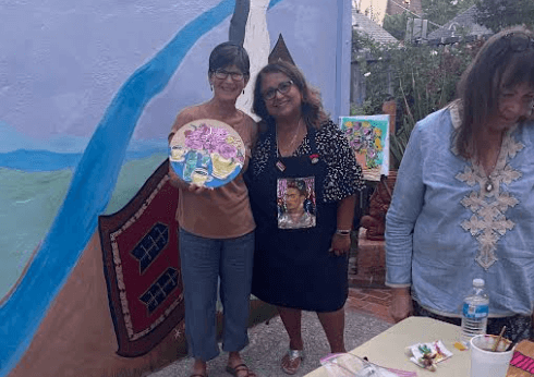 Two women standing closely next to each other in front of a colorful wall outside while one is holding a painting up in her right hand. A third female is standing in the foreground on the right edge of the frame, looking down at a table.