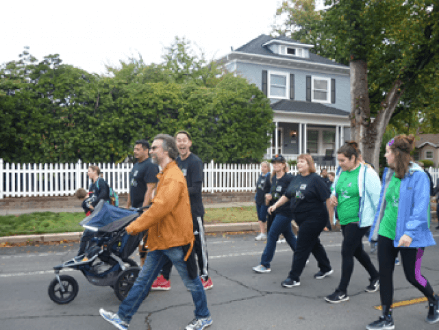 Group of men and women, one man pushing a stroller, walking in a group together down a street. Most are wearing fitness outfits.