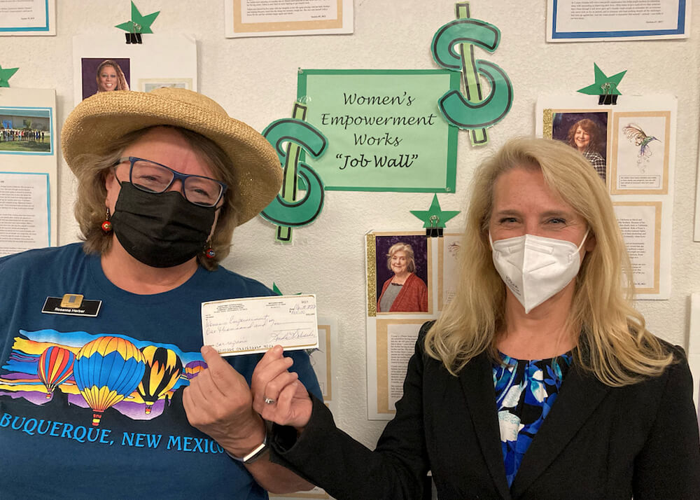 Close up of two women standing in front of a wall  with a green sign that says, "Women's Empowerment Works 'Job Wall'" while each holding up the same check.